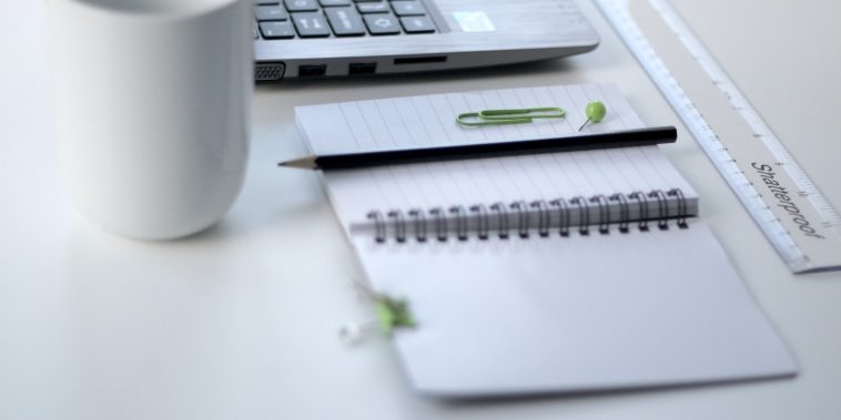 black pencil on ruled notepad beside white ceramic mug and gray laptop computer